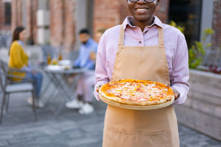 Smiling waitress serving pizza on an outdoor patio, with other patrons in the background enjoying their meals.の写真素材