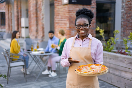 A smiling waitress holds out a pizza outdoors, inviting patrons. Diners sit at tables in the blurred background.の写真素材