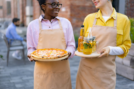 Two waitresses smiling and holding food and drinks outdoors at a restaurant.の写真素材