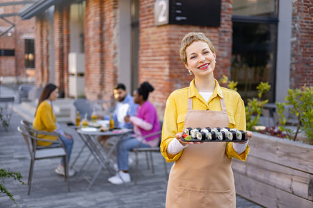 A smiling waitress presents sushi rolls outdoors on a patio. Customers dine in the background.の写真素材