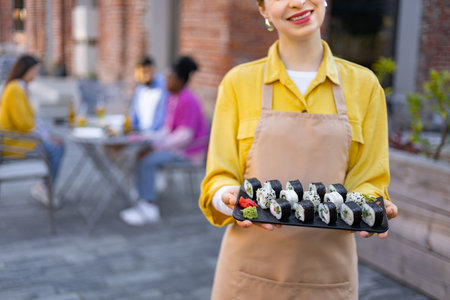 A smiling waitress holds a platter of sushi outdoors. Diners are blurred in the background, enjoying their meals.の写真素材