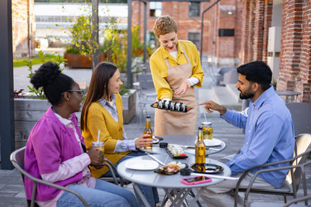 A server presents sushi to a group of friends sitting at an outdoor restaurant table, drinks and food are visible.の写真素材
