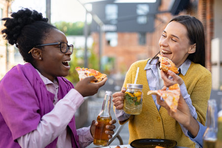 Two cheerful women share pizza and drinks outdoors, enjoying a relaxed meal together, laughing and...