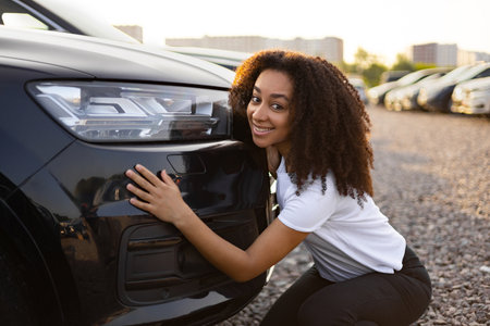 A smiling woman embraces a new black car, likely at a car dealership or lot, excited about her purchase.の写真素材