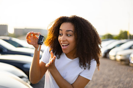 A joyful woman excitedly displays her new car keys, standing in a car dealership parking lot.の写真素材