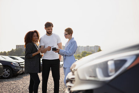 A couple is paying for a car at a dealership using a credit card, assisted by a saleswoman in a car lot.の写真素材