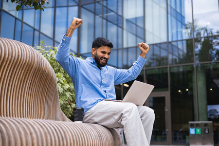 A happy man with a beard celebrates a successful project on his laptop while seated outside a modern...