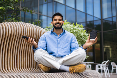 A man in a blue shirt meditates on a wooden bench, holding a coffee cup and a phone in an outdoor...