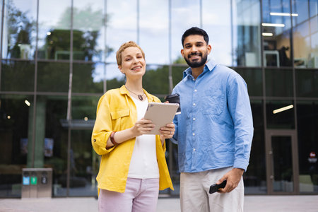Two diverse business colleagues stand in front of a modern office building, smiling and looking at the camera, one holding a tablet.の写真素材