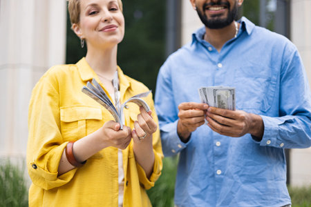 A diverse couple joyfully holds stacks of American hundred dollar bills, symbolizing financial...