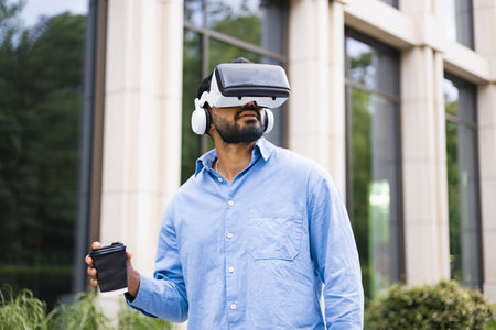 An individual wears a VR headset with headphones and holds a coffee cup, experiencing virtual...
