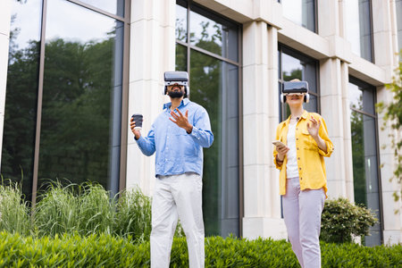 Two people wearing VR headsets explore a virtual reality experience outdoors in front of a modern...