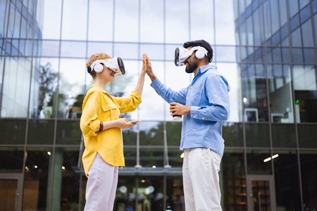 Two people wearing VR headsets give each other a high five outside a modern office building.