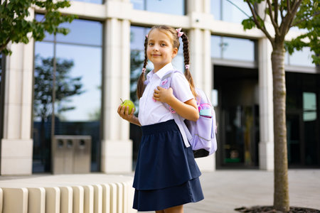A young girl with pigtails carries a backpack and an apple, smiling at the camera in front of a school building.の写真素材
