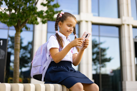 A young girl with pigtails, a backpack, and school uniform is looking at her phone while sitting in front of a school.の写真素材