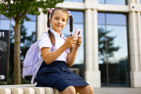 Smiling schoolgirl in uniform with backpack and smartphone, seated outside the school building.の写真素材