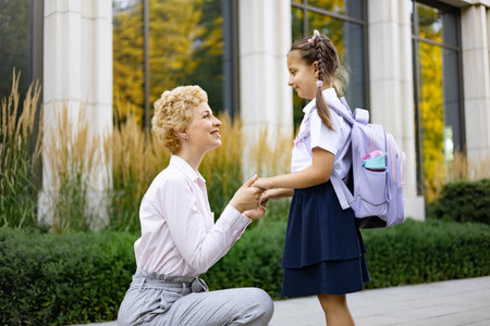 A tender moment between a mother and her young daughter at the school. The mom is kneeling, holding hands with the girl.の写真素材