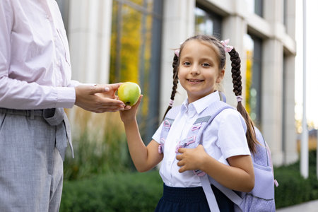 A mother offers her daughter a green apple at the schoolyard. The girl wears a backpack and uniform.の写真素材