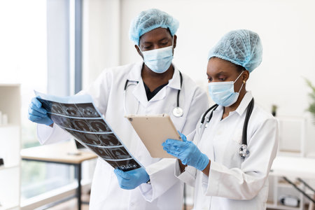 Two adult medical professionals discussing diagnostics, one viewing scans and one holding clipboard,...
