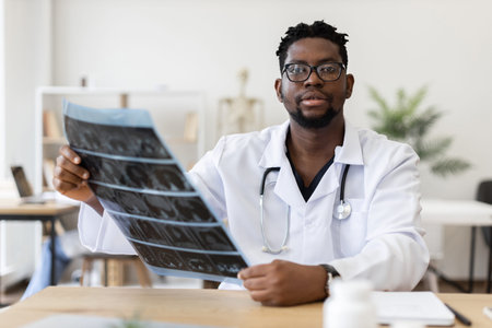 Male African doctor wearing white coat with stethoscope analyzing X-ray in modern office during...