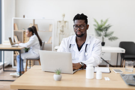 Male doctor using laptop in healthcare office while female colleague works in background. Modern medical environment with focus on technology, collaboration, and professional expertise.の写真素材