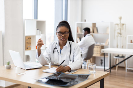 Female adult doctor wearing stethoscope writing medical notes while holding pill bottle. Bright medical office with laptop, notebook, x-rays, and colleague visible in background.の写真素材