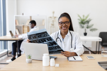 Young African American female doctor examining X-ray scan in modern medical office beside male colleague working. Represents professional healthcare, diagnosis, collaborative environment.の写真素材
