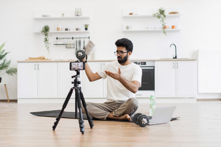 Young man sits on yoga mat in kitchen filming workout tutorial with smartphone cameraの写真素材