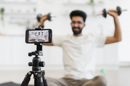 Male adult records fitness video blog using smartphone while using dumbbells for strength exercises. Seen seated in modern home interior, showing a health-focused and technology-driven lifestyle.の写真素材