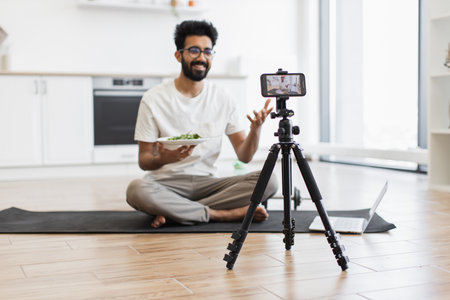 Young adult man sitting cross-legged on yoga mat in modern kitchen recording video blog with smartphone and tripod holding healthy salad plate. Scene suggests health, nutrition, video blogging.の写真素材