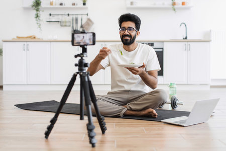 Middle age man sitting on yoga mat in bright kitchen recording healthy eating blog with smartphone...