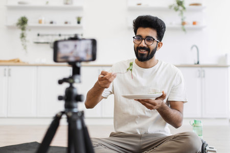 Young adult man sits in kitchen recording video about healthy eating habits. Scene shows him eating...