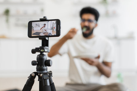 Young adult man sits in kitchen recording video about healthy eating habits. Scene shows him eating and using smartphone camera for vlog, promoting balanced diet and modern lifestyle.の写真素材