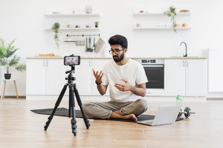 Man sitting on floor in modern living room recording informative fitness blog using smartphone. Explains exercise benefits to viewers. Promotes healthy lifestyle, well-being, engagement.の写真素材