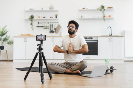 Young adult man sitting in modern kitchen recording video blog about fitness and mindfulness while meditating. Promoting healthy lifestyle including mental and physical wellness to his online audienceの写真素材