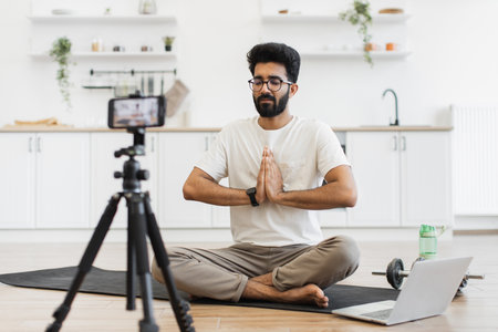 Young adult man sitting in modern kitchen recording video blog about fitness and mindfulness while meditating. Promoting healthy lifestyle including mental and physical wellness to his online audienceの写真素材