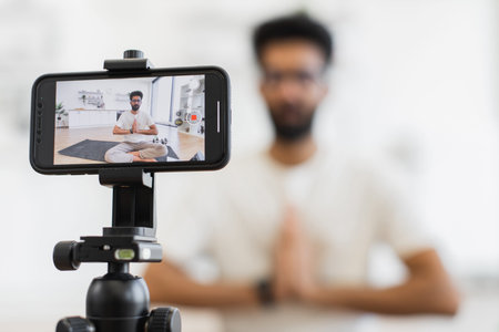 Young adult man meditating on yoga mat at home while recording his video blog focusing on fitness...
