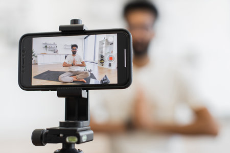 Bearded adult man sitting cross-legged on a yoga mat, filming meditation video focused on mindfulness and wellness. Modern bright home with phone on tripod for blogging or fitness tutorial.の写真素材