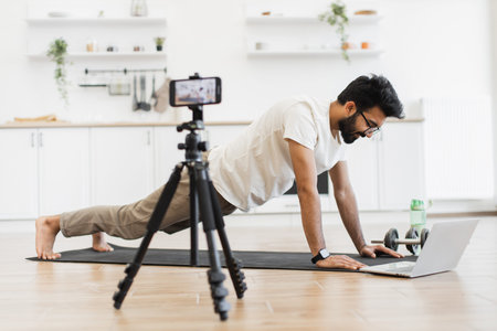 Young adult man in living room recording fitness blog video exercising on mat using laptop.