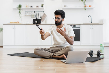 Young adult man recording video blog about fitness and health at home, sitting cross-legged on mat, using smartphone mounted on stick, with laptop and water bottle nearby in bright kitchen setting.の写真素材