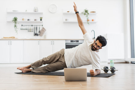 Young adult man in his 30s performs fitness side plank in bright minimalist kitchen using yoga mat,...