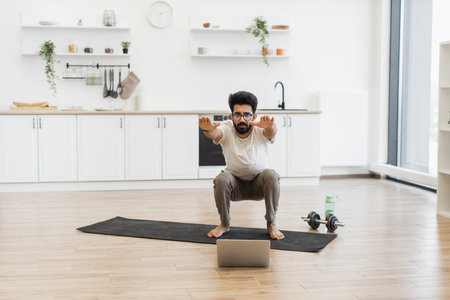 Young man performing squats in bright home kitchen using laptop for virtual fitness routine....