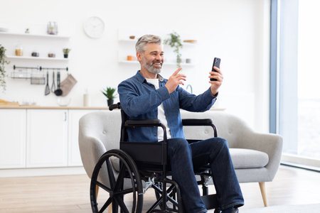 Mature Caucasian man in wheelchair using smartphone for video call in modern bright living room. Depicts inclusive technology use and connectivity.の写真素材