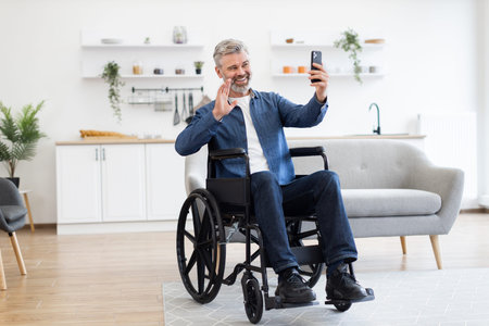 Mature Caucasian man seated in wheelchair holding smartphone waving during video call within home...