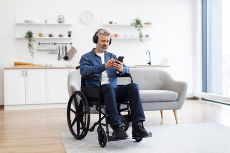 Mature Caucasian man in wheelchair enjoys music using headphones and phone in bright modern living...