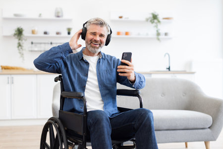 Mature Caucasian man in wheelchair wearing headphones using phone happily dancing in kitchen environmentの写真素材