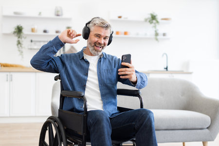 Mature Caucasian man in wheelchair wearing headphones using phone happily dancing in kitchen environmentの写真素材