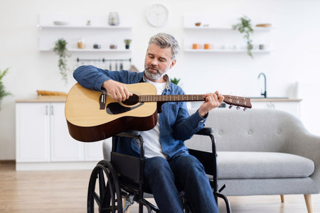 Bearded middle-aged man in wheelchair playing acoustic guitar indoors. Casual home environment...