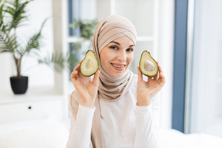 Smiling young Muslim woman holding avocado halves, wearing hijab in modern bright interior setting