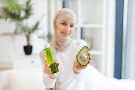 Focused view on Muslim adult woman with turban in modern room holding half avocado and green...